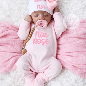 May include: A light pink long-sleeved footed onesie with the text Nia Rose in pink script, paired with a white beanie featuring a large pink bow and the name Nia. A light pink pacifier is attached to a wooden bead clip chain.