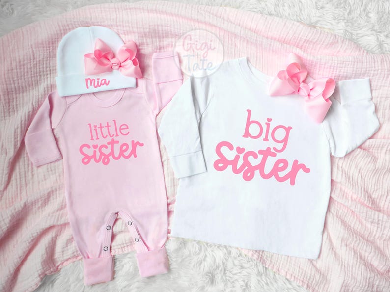 May include: A light pink long-sleeve onesie with the text little sister in pink, paired with a white beanie featuring a pink bow and the name Mia. Beside it, a white long-sleeve shirt with the text big sister in pink and a pink bow.