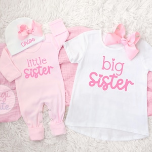 May include: A light pink long-sleeve romper with the text little sister, a white beanie with a pink bow and the name Chloe, and a white short-sleeve t-shirt with the text big sister and a matching pink bow.