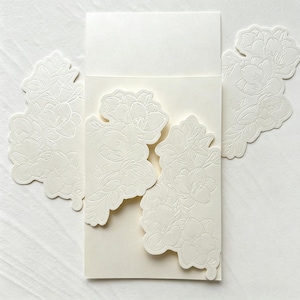 May include: A cream-colored cardstock invitation folder featuring intricate, embossed floral patterns on the front flaps. The design includes detailed outlines of flowers and leaves, with a smooth, blank rectangular space at the top for text.