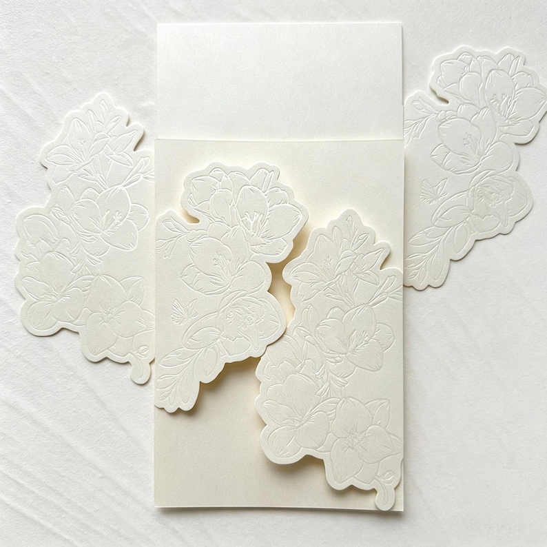 May include: A cream-colored cardstock invitation folder featuring intricate, embossed floral patterns on the front flaps. The design includes detailed outlines of flowers and leaves, with a smooth, blank rectangular space at the top for text.