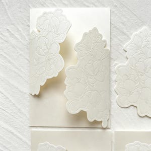 May include: Off-white rectangular cardstock stationery featuring intricate, raised floral embossing on the front flaps. The design showcases detailed botanical patterns with petals and leaves, creating a textured, monochromatic appearance.