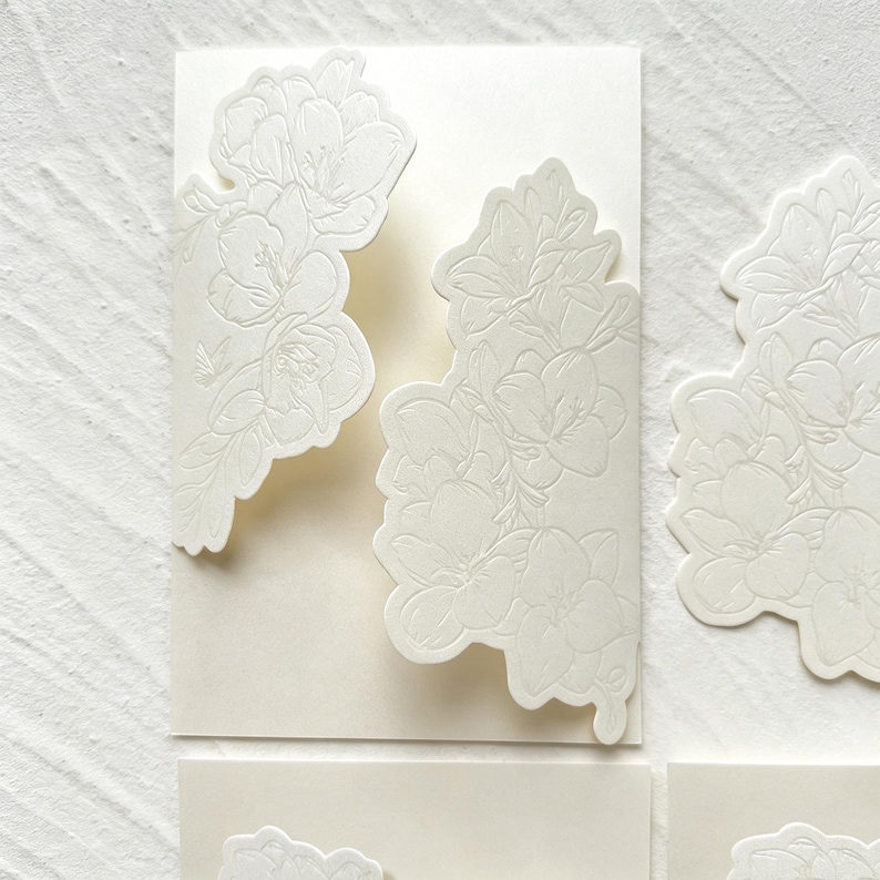 May include: Off-white rectangular cardstock stationery featuring intricate, raised floral embossing on the front flaps. The design showcases detailed botanical patterns with petals and leaves, creating a textured, monochromatic appearance.