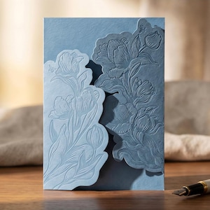 Könnte beinhalten: Eine hellblaue Einladungskarte mit floralem Design. Die Karte hat ein geprägtes Tulpenmuster auf der Vorderseite mit einem gestanzten Rand. Die Karte besteht aus strukturiertem Papier und eignet sich für Hochzeiten oder besondere Anlässe.