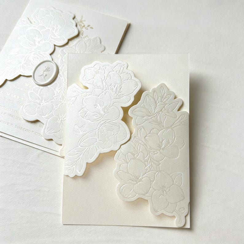 May include: Two cream-colored invitation cards featuring intricate, raised floral embossing on the front panels. The cards have die-cut edges that follow the floral outlines, and one card includes a small, oval-shaped wax seal with a floral motif.