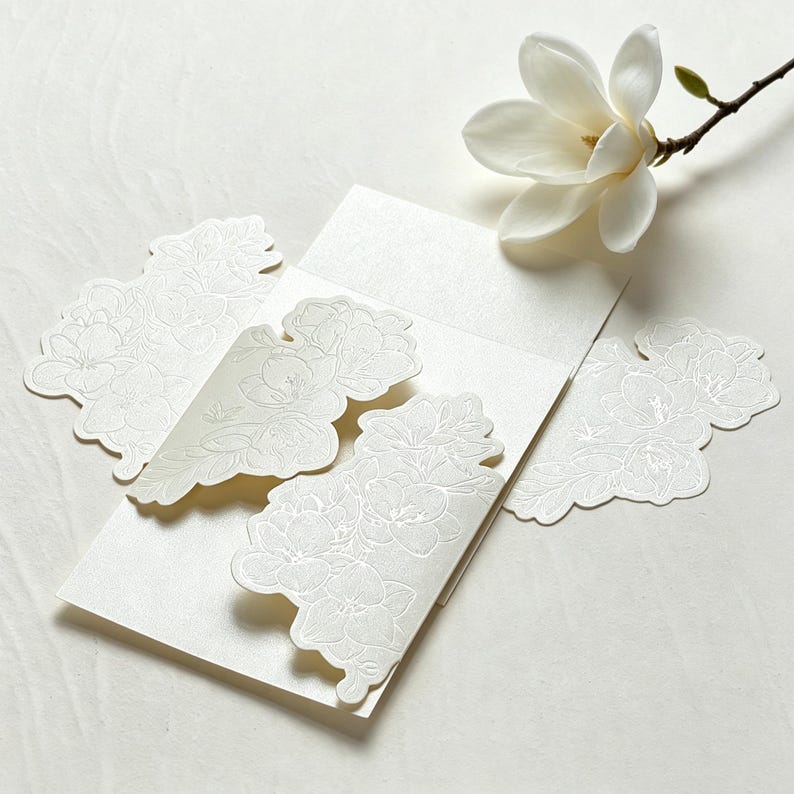 May include: A set of cream-colored stationery featuring intricate embossed floral patterns on the edges. The paper has a subtle pearlescent sheen and is accompanied by a single fresh white magnolia flower with a brown stem and green bud.