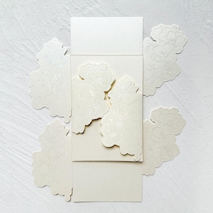 May include: A cream-colored paper card set featuring a central rectangular panel with die-cut floral edges that overlap when closed. The paper has a subtle shimmer and is decorated with delicate, embossed line-art illustrations of blooming flowers.