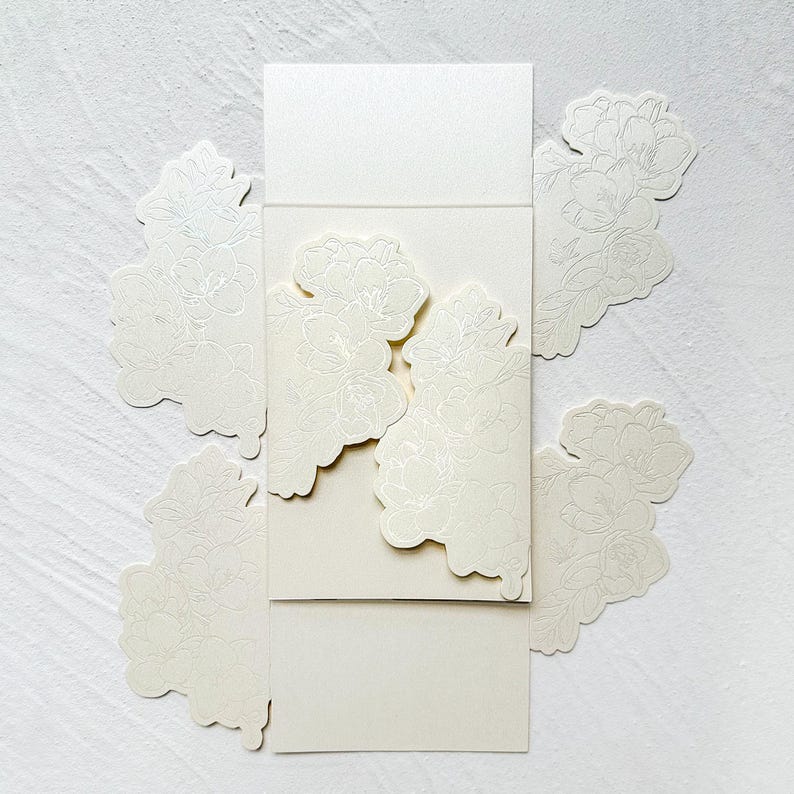 May include: A cream-colored paper card set featuring a central rectangular panel with die-cut floral edges that overlap when closed. The paper has a subtle shimmer and is decorated with delicate, embossed line-art illustrations of blooming flowers.