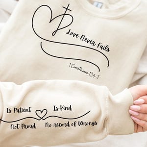 Op de afbeelding: Crèmekleurige sweatshirt met zwart schrift. De voorkant heeft een hart- en kruisontwerp met de woorden "Love Never Fails" en "1 Korintiërs 13:4-7." De mouw toont "Is Patient Is Kind Not Proud No record of Wrongs."