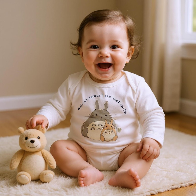 May include: A smiling baby wearing a white long-sleeved onesie with a cartoon illustration of two characters and the text "Made on eardust and soot sprites." A stuffed teddy bear is next to the baby.