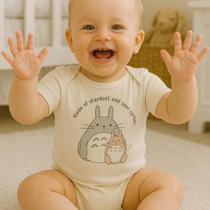 May include: A smiling baby wearing a cream-colored onesie with a graphic of two cartoon characters and the text "Made of stardust and sool sprites." The baby is sitting on a white rug with arms raised.