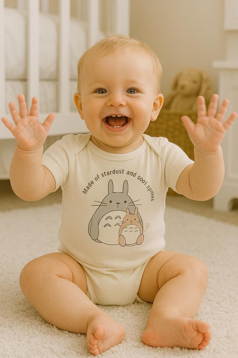 May include: A smiling baby wearing a cream-colored onesie with a graphic of two cartoon characters and the text "Made of stardust and sool sprites." The baby is sitting on a white rug with arms raised.