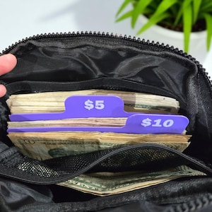 Op de afbeelding: Zwarte etui met ritssluiting en paarse verdelers met de tekst '$5' en '$10', die stapels papiergeld scheiden. De etui is open en onthult de inhoud. Een hand is zichtbaar aan de linkerkant van de afbeelding.