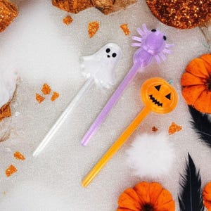 Könnte beinhalten: Ein Halloween-Bild mit drei dekorativen Getränkerührern: einem weißen Geist, einer lila Spinne und einem orangefarbenen Kürbis. Die Rührer sind vor einem Hintergrund aus orangefarbenem und goldenem Glitzer, mit orangefarbenen Filzkürbissen und schwarzen Federn.