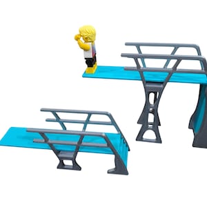 Puede incluir: Juego de trampolín de dos piezas con plataforma azul claro y soportes grises. Una figura amarilla con una camisa colorida está de pie en una de las tablas, con los brazos levantados. El juego está diseñado para jugar.