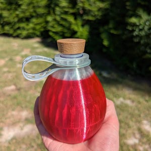 May include: A red, teardrop-shaped bottle with a brown cork and a clear plastic loop. The bottle is filled with a red liquid. The bottle is held in a hand against a background of green foliage.