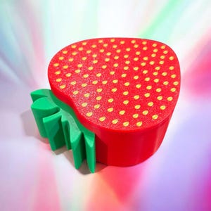 May include: A red and green strawberry-shaped trinket box with a gold glitter finish. The box has a detailed design of small yellow seeds on the top.