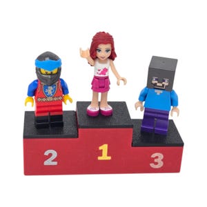 May include: A red podium with black steps displays three Lego figures. The figures include a knight, a girl with red hair, and a Minecraft character. The podium has the numbers 1, 2, and 3 in yellow and white.