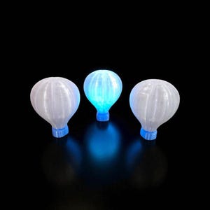 Puede incluir: Tres luces decorativas de globo aerostático. Dos son blancas y una brilla con una luz azul. Cada globo tiene una base azul. El fondo es negro.