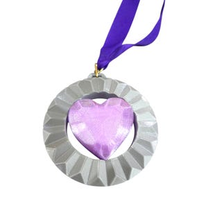 May include: A gray, geometric-patterned ornament with a purple heart-shaped center. The ornament is suspended by a purple ribbon. The heart has a faceted surface, and the ornament is likely for decoration.