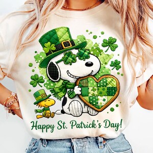 Puede incluir: Camiseta color crema con un diseño del Día de San Patricio. El gráfico incluye a Snoopy con un sombrero y bufanda verdes, sosteniendo un corazón, y el texto "Happy St. Patrick's Day!" en verde.
