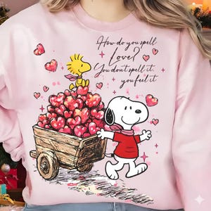 Puede incluir: Sudadera rosa con Snoopy y Woodstock. Snoopy tira de un carro lleno de corazones. El texto dice: "¿Cómo se escribe Amor? No se escribe, se siente". Corazones y estrellas dispersos.