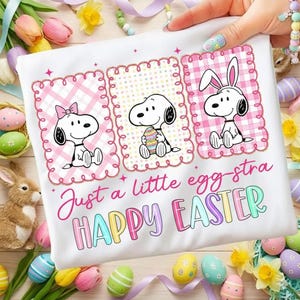 Puede incluir: Camiseta blanca con ilustraciones de Snoopy en paneles enmarcados en rosa, celebrando la Pascua. Un Snoopy lleva un lazo, otro sostiene un huevo y el último tiene orejas de conejo. Texto: "Just a little egg-stra HAPPY EASTER."