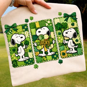 Puede incluir: Sudadera beige con tres diseños de patchwork verdes con Snoopy, tréboles y lazos. Un Snoopy sostiene un trébol, otro tiene un trébol con la palabra "LUCKY", y el tercero tiene un lazo de trébol. El diseño es temático del Día de San Patricio.