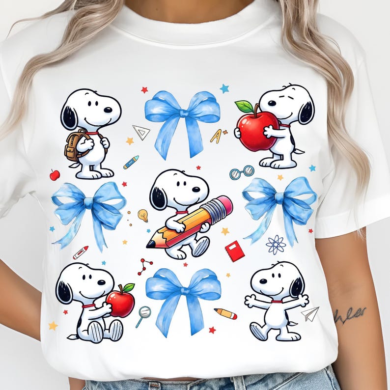 May include: White t-shirt featuring cartoon Snoopy characters with school-themed graphics. The design includes Snoopy with a backpack, holding an apple, and with a pencil. Blue bows, red apples, and school supplies are also featured.