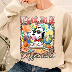 Puede incluir: Sudadera beige con un gráfico de Snoopy con gafas de sol y soplando una burbuja. El gráfico incluye el texto "It's OK to be Different" y salpicaduras de pintura coloridas. Un globo terráqueo, libros y piezas de rompecabezas también están presentes.