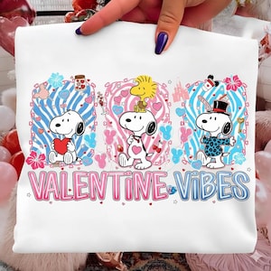 May include: White t-shirt featuring Snoopy characters with Valentine's Day graphics. The design includes hearts, flowers, and the text "VALENTINE VIBES" in pink and blue. The shirt has a crew neck.