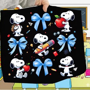 May include: Black blanket featuring Snoopy characters with school-themed elements. The design includes Snoopy with a backpack, holding an apple, and a pencil, along with blue bows, stars, and school supplies. The blanket is held up by a person.