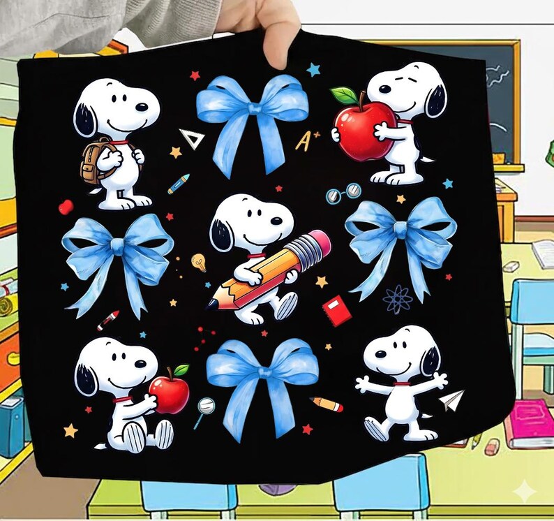 May include: Black blanket featuring Snoopy characters with school-themed elements. The design includes Snoopy with a backpack, holding an apple, and a pencil, along with blue bows, stars, and school supplies. The blanket is held up by a person.