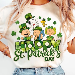 Puede incluir: Camiseta color crema con un diseño del Día de San Patricio. El gráfico incluye personajes de dibujos animados con sombreros y trajes verdes, con las palabras "HAPPY St. Patrick's DAY" en letras verdes. Acentos de trébol están dispersos alrededor del diseño.