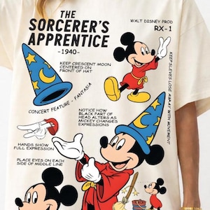 Puede incluir: Camiseta color crema con un gráfico de Mickey Mouse de "El aprendiz de brujo". El diseño incluye a Mickey con un sombrero de mago, texto e ilustraciones de la película de 1940. La camiseta tiene mangas cortas.
