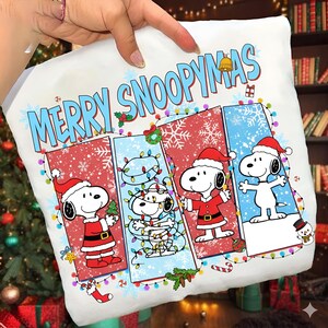 May include: White cushion featuring Snoopy in Santa outfits, with the words "MERRY SNOOPYMAS" in blue. The design includes Christmas lights, snowflakes, and festive holiday elements.