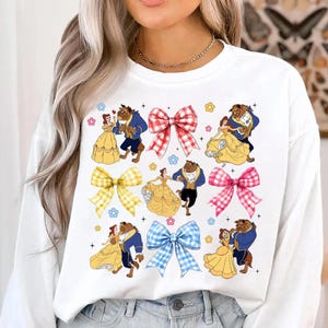 Puede incluir: Sudadera blanca con un diseño colorido que presenta ilustraciones de Bella y la Bestia bailando, intercaladas con lazos a cuadros y detalles florales. El diseño incluye elementos amarillos, azules, rojos y rosas.