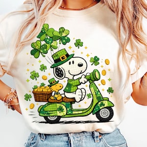 Puede incluir: Camiseta color crema con un diseño temático del Día de San Patricio. Snoopy, con sombrero y camisa verdes, monta una moto verde con Woodstock. El diseño incluye tréboles, monedas de oro y estrellas.