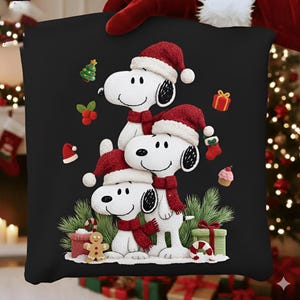 Puede incluir: Cojín cuadrado negro con un diseño festivo de Snoopy. Tres personajes de Snoopy están apilados, usando gorros de Papá Noel y bufandas rojas. Los adornos de árbol de Navidad, cajas de regalo y hombre de jengibre completan el tema navideño.