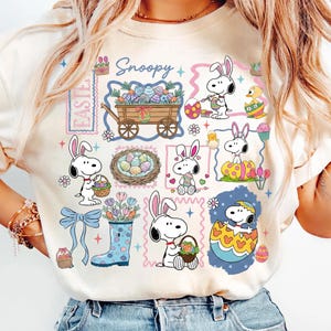 Puede incluir: Camiseta color crema con gráficos de Snoopy y temática de Pascua. El diseño incluye personajes de Snoopy con huevos de Pascua, cestas y un carro lleno de huevos decorados. La palabra "Snoopy" está escrita en cursiva.