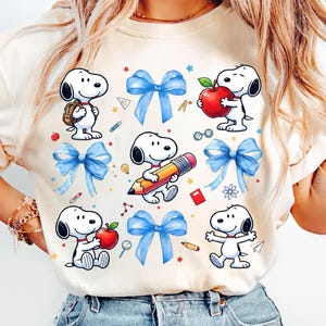 May include: A cream-colored t-shirt featuring a repeating pattern of Snoopy characters, blue bows, red apples, pencils, and school-themed graphics. The design is playful and colorful, perfect for back-to-school or everyday wear.