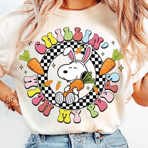 Op de afbeelding: Crèmekleurig T-shirt met een cartoonkonijn dat een wortel vasthoudt, omringd door wortels en de tekst "Chillin' With My Peeps" in een kleurrijk retro-lettertype. Het ontwerp bevat een zwart-wit geblokte cirkel.