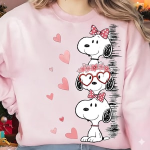 Puede incluir: Sudadera rosa claro con un diseño de Snoopy. Tres personajes de Snoopy apilados, cada uno con un accesorio diferente: un lazo, gafas en forma de corazón y una corona floral. Corazones rosas dispersos.