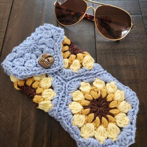 May include: A crocheted sunglasses case with a blue background and a sunflower pattern in yellow, brown, and cream. The case has a flap with a button closure.