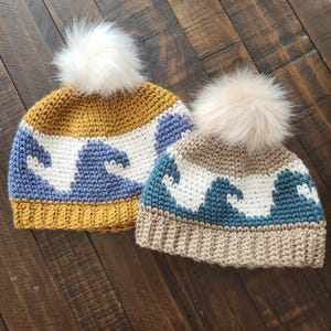 May include: Two crocheted hats with wave patterns and faux fur pom poms. The hats are in different color combinations: one is mustard yellow, white, and blue, and the other is beige, white, and teal.