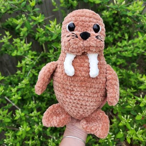 May include: A handmade, plush walrus toy in a soft, muted brown color. The walrus has large, black eyes, a black nose, and white tusks. The toy is crocheted with a textured yarn, giving it a cuddly appearance. The background is a lush green bush.