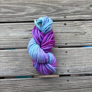 May include: A skein of chunky yarn in shades of blue, purple, and magenta. The yarn is thick and appears soft, with a gradient of colors. The yarn is resting on a weathered wooden surface.