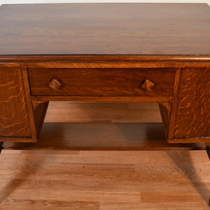 May include: A wooden desk with a dark brown finish. The desk has a rectangular top, a single drawer with diamond-shaped pulls, and two side cabinets. The desk is supported by four legs and has a shelf below the drawer.