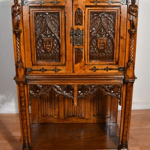 May include: A dark wood cabinet with intricate carvings and a Gothic design. The cabinet features two doors with detailed relief panels, decorative metal hardware, and carved figures on the sides. The lower section has an open shelf.