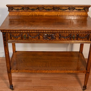 May include: A wooden antique table with a shelf and drawer. The table is made of a light brown wood with a decorative trim around the drawer and top. The table has four legs with small wheels.